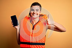 Young beautiful brunette woman wearing safe lifejacket showing smartphone happy with big smile doing ok sign, thumb up with