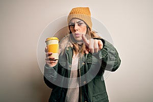 Young beautiful brunette woman wearing snow clothes drinking cup of coffee pointing with finger to the camera and to you, hand