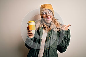 Young beautiful brunette woman wearing snow clothes drinking cup of coffee pointing and showing with thumb up to the side with