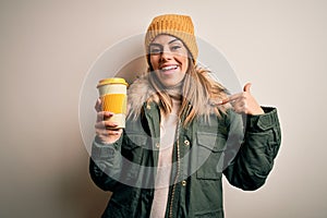 Young beautiful brunette woman wearing snow clothes drinking cup of coffee with surprise face pointing finger to himself