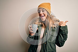 Young beautiful brunette woman wearing snow clothes drinking mug of coffee pointing and showing with thumb up to the side with