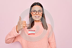 Young beautiful brunette woman wearing sticker with hello my name is message Smiling happy and positive, thumb up doing excellent
