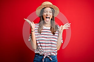 Young beautiful brunette woman wearing striped t-shirt and summer hat over red background celebrating mad and crazy for success