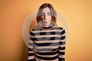 Young beautiful brunette woman wearing striped turtleneck sweater over yellow background afraid and shocked with surprise