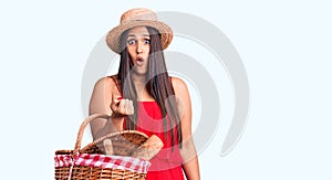 Young beautiful brunette woman wearing summer hat holding picnic basket scared and amazed with open mouth for surprise, disbelief