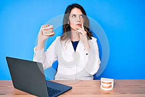 Young beautiful brunette woman working at dentist clinic holding denture serious face thinking about question with hand on chin,