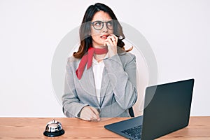 Young beautiful brunette woman working at hotel reception using laptop looking stressed and nervous with hands on mouth biting