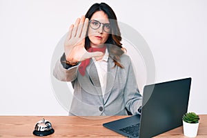 Young beautiful brunette woman working at hotel reception using laptop with open hand doing stop sign with serious and confident