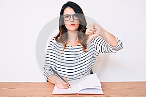 Young beautiful brunette woman writing book sitting on the table with angry face, negative sign showing dislike with thumbs down,