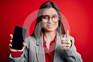 Young beautiful business woman with blue eyes holding smartphone showing screen happy with big smile doing ok sign, thumb up with