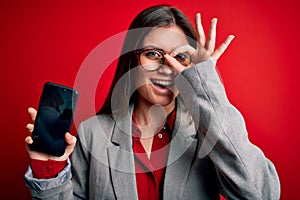 Young beautiful business woman with blue eyes holding smartphone showing screen with happy face smiling doing ok sign with hand on