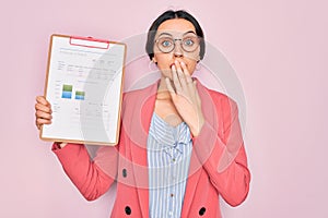 Young beautiful business woman with blue eyes wearing glasses holding clipboard cover mouth with hand shocked with shame for