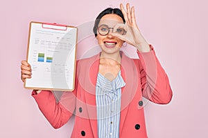 Young beautiful business woman with blue eyes wearing glasses holding clipboard with happy face smiling doing ok sign with hand on