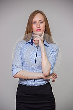 Young beautiful business woman in a blue shirt stands with her arms crossed and thinks over a gray background