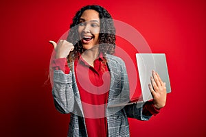 Young beautiful business woman with curly hair working using laptop over red background pointing and showing with thumb up to the