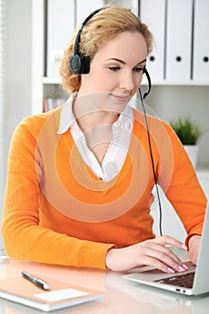 Young beautiful business woman in headset or call operator communicates by laptop computer. Orange sweater is uniform of