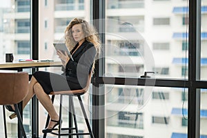 Young beautiful business woman near window using tablet in office. Smiling sexy lady confident in black suited