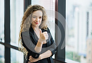 Young beautiful business woman standing near window in office. Smiling sexy lady confident in black suited showing thumbs up