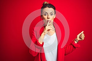 Young beautiful business woman standing over red isolated background asking to be quiet with finger on lips pointing with hand to