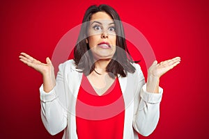 Young beautiful business woman wearing elegant jacket over red isolated background clueless and confused expression with arms and