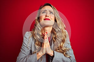 Young beautiful business woman wearing elegant jacket standing over red  background begging and praying with hands