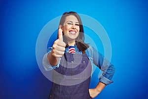 Young beautiful business woman wearing store uniform apron over blue isolated background doing happy thumbs up gesture with hand