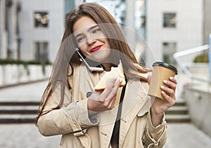 Young beautiful businesslady having phone conversation walking downthe street with cup of coffee eats sandwich on the go in a