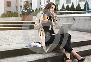 Young beautiful businesswoman bites sandwich and drinks hot coffee in paper cup works in her laptop sitting on stairs outside