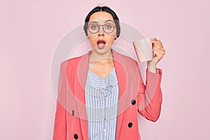 Young beautiful businesswoman with blue eyes wearing glasses drinking pink mug of coffee scared in shock with a surprise face,