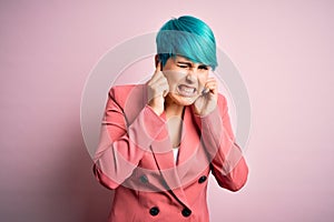 Young beautiful businesswoman with blue fashion hair wearing jacket over pink background covering ears with fingers with annoyed