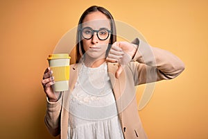 Young beautiful businesswoman drinking cup of takeaway coffee over yellow background with angry face, negative sign showing