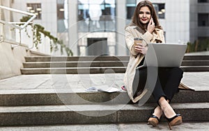 Young beautiful businesswoman having phone conversation works in her laptop drinks coffee sitting on stairs near office center in