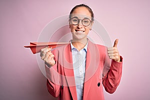 Young beautiful businesswoman holding paper airplane over isolated pink background happy with big smile doing ok sign, thumb up