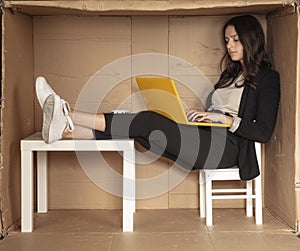 Young beautiful businesswoman sitting in office cardboard box, legs on desk and working on computer
