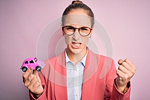 Young beautiful businesswoman wearing glasses holding pink car toy over isolated background annoyed and frustrated shouting with
