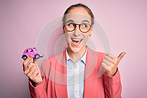 Young beautiful businesswoman wearing glasses holding pink car toy over isolated background pointing and showing with thumb up to