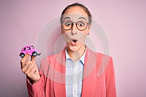 Young beautiful businesswoman wearing glasses holding pink car toy over isolated background scared in shock with a surprise face,
