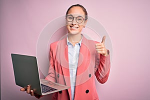 Young beautiful businesswoman wearing glasses working using laptop over pink background happy with big smile doing ok sign, thumb