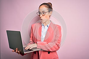 Young beautiful businesswoman wearing glasses working using laptop over pink background scared in shock with a surprise face,