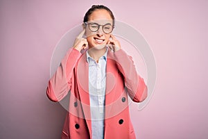 Young beautiful businesswoman wearing jacket and glasses over isolated pink background covering ears with fingers with annoyed