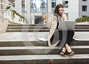 Young beautiful businesswoman works in her laptop drinks coffee sitting on stairs near office center in downtown, multitasking