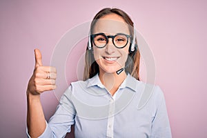 Young beautiful call center agent woman wearing glasses working using headset doing happy thumbs up gesture with hand