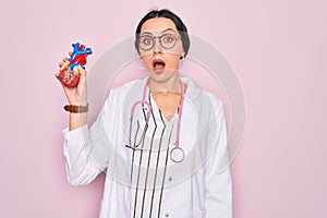 Young beautiful cardiologist woman with blue eyes wearing stethoscope holding heart scared in shock with a surprise face, afraid