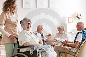 Young beautiful caregiver and positive senior woman sitting at wheelchair in nursing home for elderly