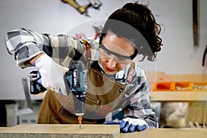 Young beautiful carpenter woman in apron holding hand drill, working with wood plank, female craft worker making wooden furniture