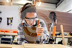 Young beautiful carpenter woman wearing safety glasses goggles and apron, using carpentry tools with wood plank to make wooden