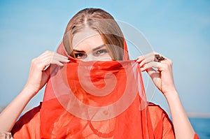 Young beautiful caucasian blond woman, with makeup, in light dress and red thin scarf on head and face against blue sky