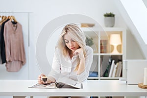 A young beautiful Caucasian blonde woman rests at home. A woman sits at a table, drinking coffee from a glass cup and