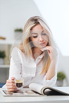 A young beautiful Caucasian blonde woman rests at home. A woman sits at a table, drinking coffee from a glass cup and