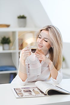 A young beautiful Caucasian blonde woman rests at home. A woman sits at a table, drinking coffee from a glass cup and